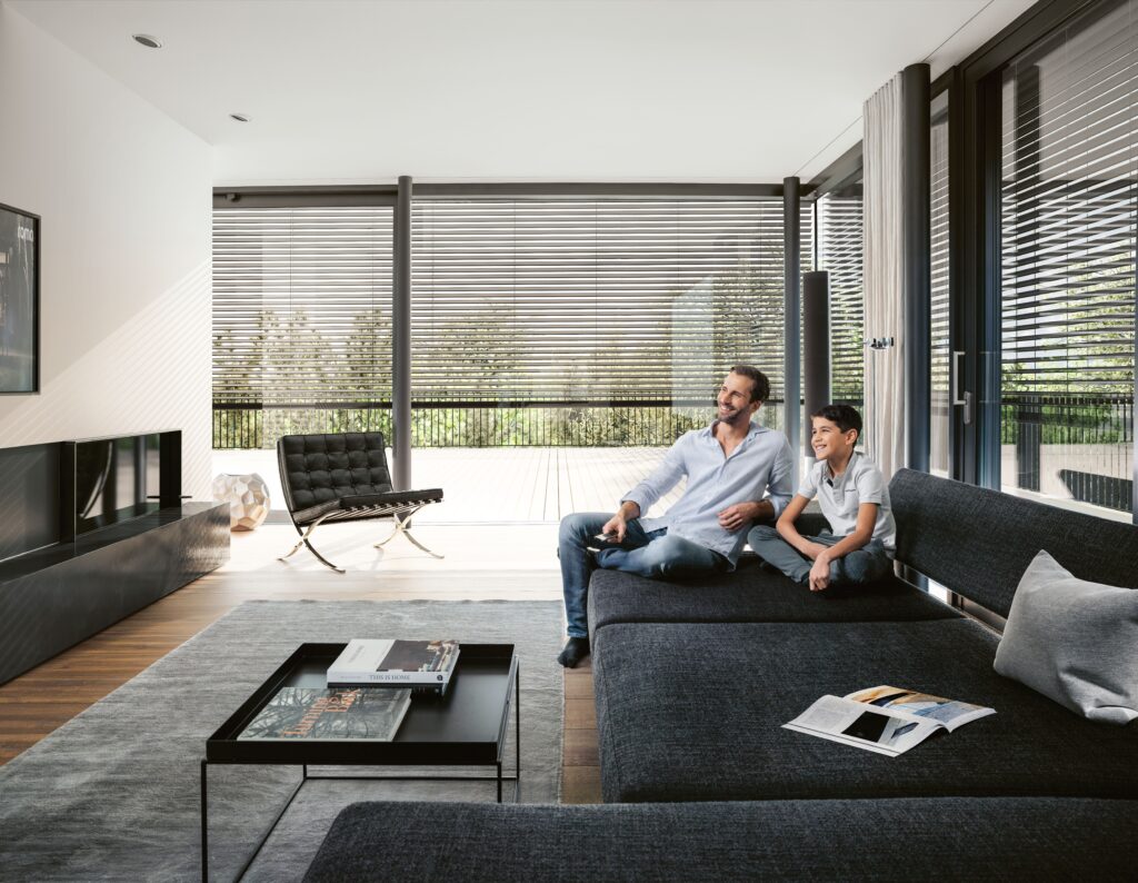 Ein Mann und ein Junge sitzen lächelnd im Wohnzimmer mit Jalousien im Hintergrund